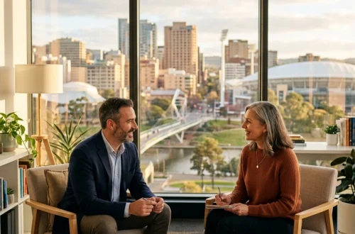 Two professionals sit in a bright office discussing, with notebooks and a tablet on the table and a cityscape outside the window.