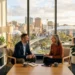 Two professionals sit in a bright office discussing, with notebooks and a tablet on the table and a cityscape outside the window.