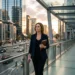 Professional woman in a navy blazer walks along a glass walkway in a modern city at sunset, carrying a brown notebook.