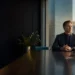 Man in a dark suit sits at a polished conference table by a window overlooking a city skyline at sunset, hands folded.
