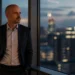 Man in a dark suit stands by a window in a high-rise office, gazing thoughtfully at the city lights at dusk.