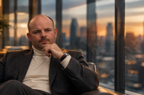 Man seated in a modern office lounge, hand on chin, gazing thoughtfully with city skyline at sunset behind glass.