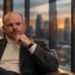 Man seated in a modern office lounge, hand on chin, gazing thoughtfully with city skyline at sunset behind glass.